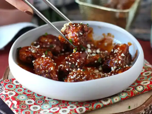 Chrupiący kurczak gochujang w sosie, posypany sezamem i orzeszkami, podany w białej misce.