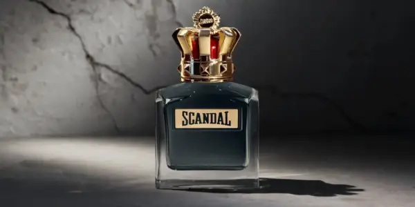 Elegancka butelka perfum Scandal męskie z koroną na nakrętce, stojąca na tle pękniętej ściany.