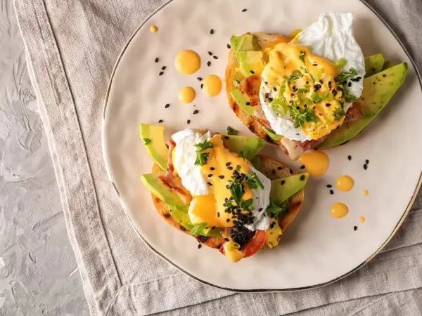 Två läckra eggs benedict på rostat bröd med avokado, bacon och hollandaisesås. Garnerade med persilja och svarta sesamfrön.