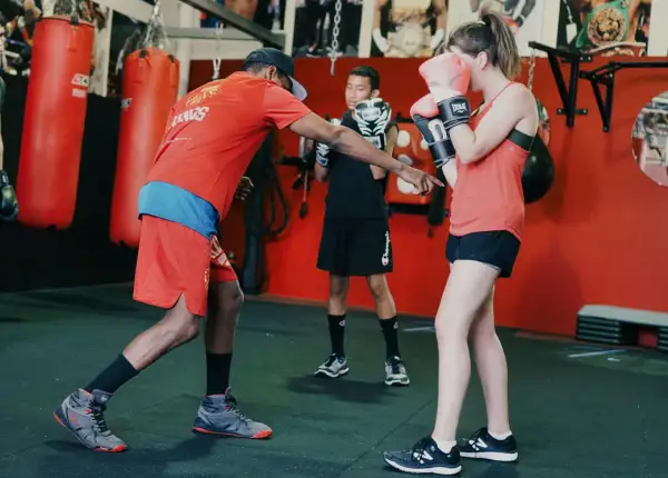 A boxing coach instructs a young woman on a proper duck punch. Another young person watches.