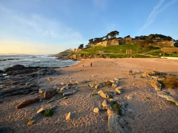 Atardecer en la playa concheira de Baiona. Olas rompen en la costa rocosa, mientras una fortaleza histórica corona la colina verde.