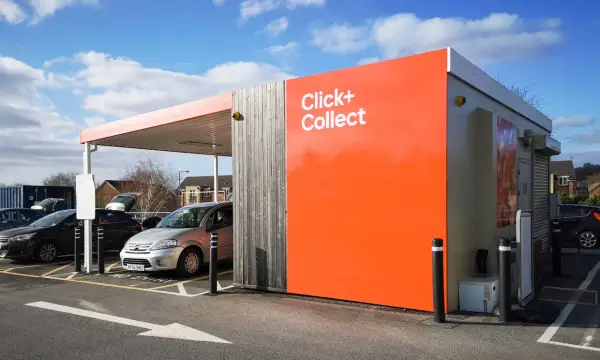 Zona de **click and collect** con coches aparcados bajo un toldo. Edificio naranja brillante con el texto "Click+ Collect".