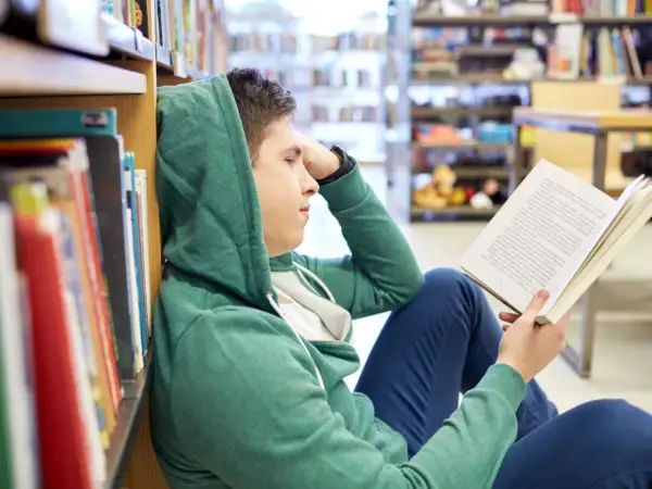 Chłopak w kapturze czyta książkę, szukając streszczenia makbeta. Siedzi na podłodze w bibliotece, opierając się o regał.