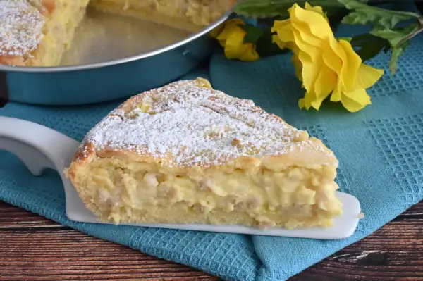 Una fetta di pastiera napoletana con crema pasticcera, cosparsa di zucchero a velo, su un piatto bianco, con fiori gialli sullo sfondo.