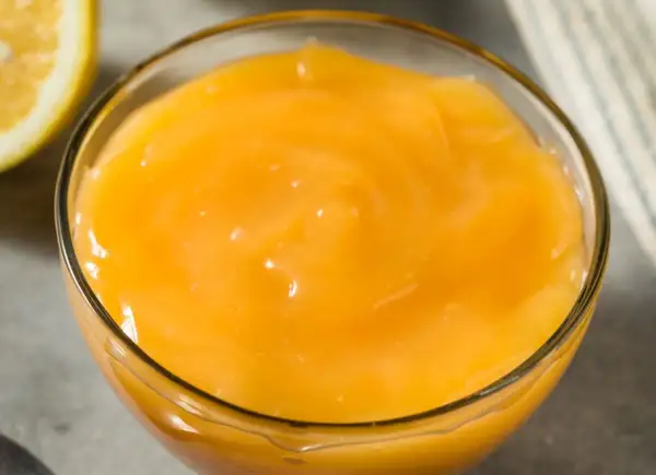 Gładki, żółty lemon curd w szklanej miseczce, obok połówka cytryny. Idealny do deserów.