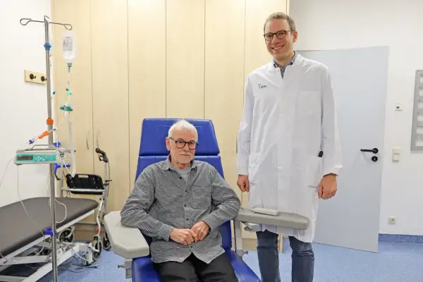 Ein Arzt steht neben einem Patienten, der für eine Alzheimer Behandlung in einem Behandlungsstuhl sitzt. Infusionsständer und medizinische Geräte sind im Hintergrund sichtbar.