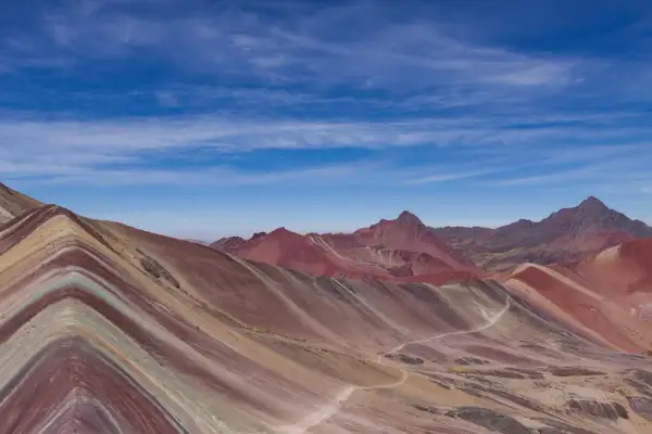 La montagne Vinicunca, aux couleurs vives, s'étend sous un ciel bleu parsemé de nuages. Un sentier serpente à travers ses flancs striés.