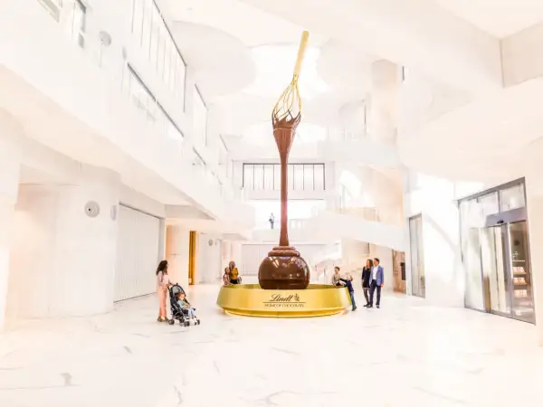 Imponerande chokladfontän i Lindt Home of Chocolate-museet. Familjer beundrar den gigantiska skulpturen i den ljusa, moderna byggnaden.
