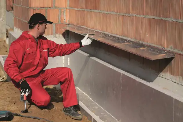 Pracownik w czerwonym kombinezonie montuje okapnik na ścianie zewnętrznej, przygotowując ją do montażu wełny mineralnej.