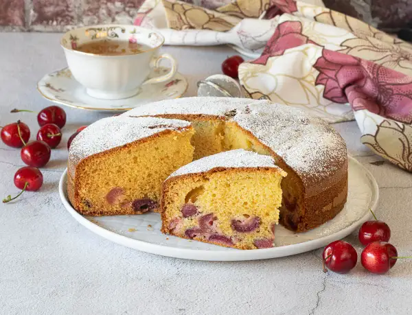 Una fetta di torta alle ciliegie, soffice e profumata, cosparsa di zucchero a velo. Ciliegie fresche e una tazza di tè completano la scena.