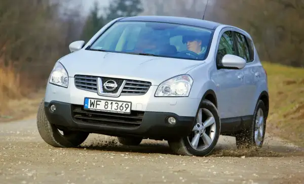 Srebrny Nissan Qashqai 1. pokonuje zakręt na piaszczystej drodze, wzbijając kurz.