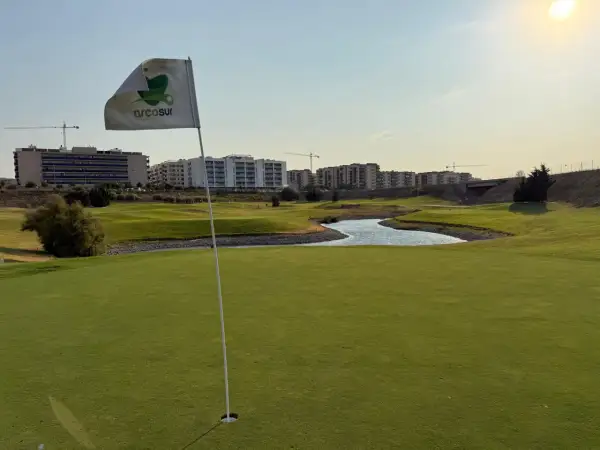 Bandera de Arcosur en un campo de golf, con edificios residenciales y grúas de construcción al fondo.