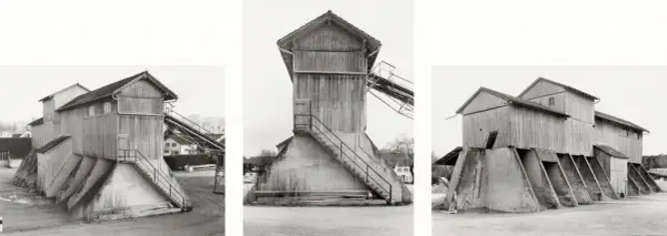 Strutture industriali in legno e cemento, tipiche delle opere di Hilla Becher, con scale esterne e nastri trasportatori.