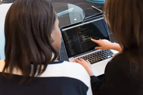 Dwie kobiety analizują kod na laptopie. Jedna wskazuje palcem na ekran, druga pisze. Tak wygląda praca programisty.