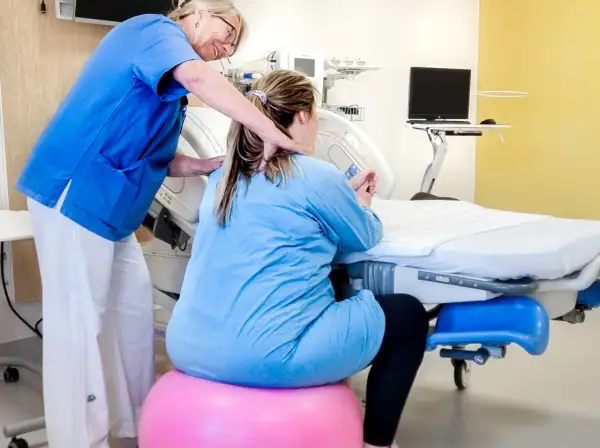 Barnmorska stöttar en kvinna i gravid vecka 36 som sitter på en rosa boll vid en förlossningssäng.