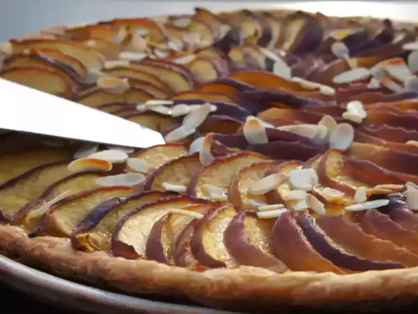 Une tranche de tarte aux nectarines, garnie d'amandes effilées, est coupée par un couteau.