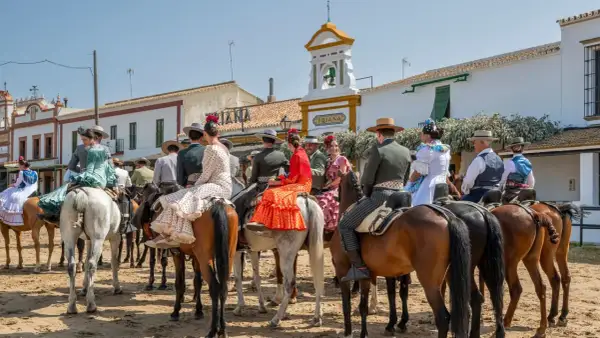 Gente vestida de flamenco a caballo, en un **camino rocío** bajo un cielo azul. Edificios blancos y un campanario al fondo.