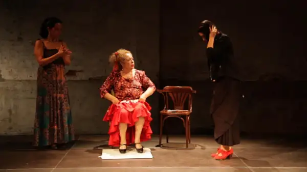 Tres mujeres en escena. Una sentada, con falda roja y blusa estampada, como **la Chana**. Otra de pie, con vestido largo y estampado. La tercera, de negro, con el pelo recogido.
