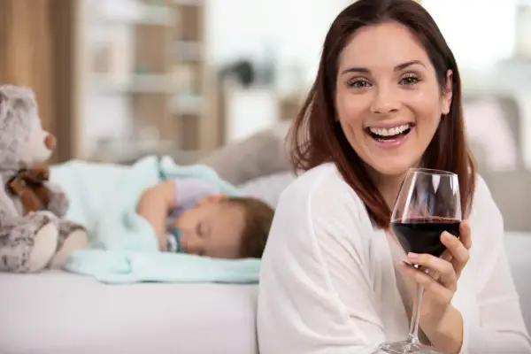 Uśmiechnięta kobieta z kieliszkiem czerwonego wina obok śpiącego dziecka. Czy można pić alkohol przy kamicy żółciowej?