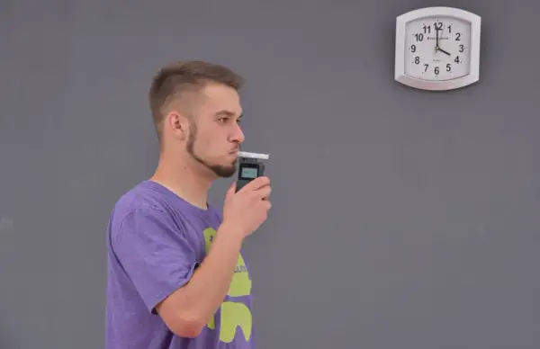 Młody mężczyzna dmucha w alkomat, sprawdzając, ile promili ma po lampce szampana. Zegar na ścianie pokazuje czas.