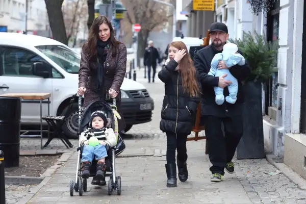 Rodzina spaceruje po mieście, mama pcha wózek z niemowlakiem, a tata niesie drugie dziecko. Wszyscy liroy dzieci wyglądają na szczęśliwych.
