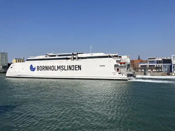 Biały prom "Bornholmslinjen Express" wypływa z portu, gotowy na podróż do Szwecji. W tle widać zabudowania portowe.