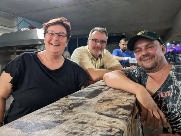 Fröhliche Menschen am holzmarkt ilsfeld. Eine Frau mit Brille und schwarzem Shirt, ein Mann mit gelbem Hemd und ein Mann mit Kappe lächeln in die Kamera.