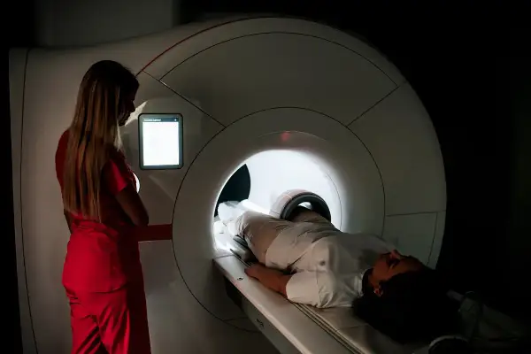 Pacjentka leży w aparacie do rezonansu magnetycznego. Technik w czerwonym stroju nadzoruje badanie. Sprawdź rezonans magnetyczny - cennik.