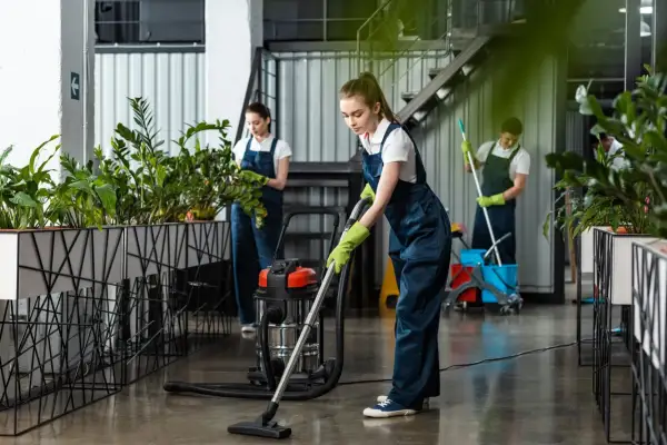 Dziewczyna w granatowym stroju do sprzątania odkurza podłogę. W tle inni pracownicy dbają o czystość i zieleń.