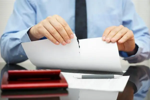 Uomo in camicia blu strappa un documento, simbolo di annullamento proposta di acquisto immobile. Calcolatrice e penna sul tavolo.