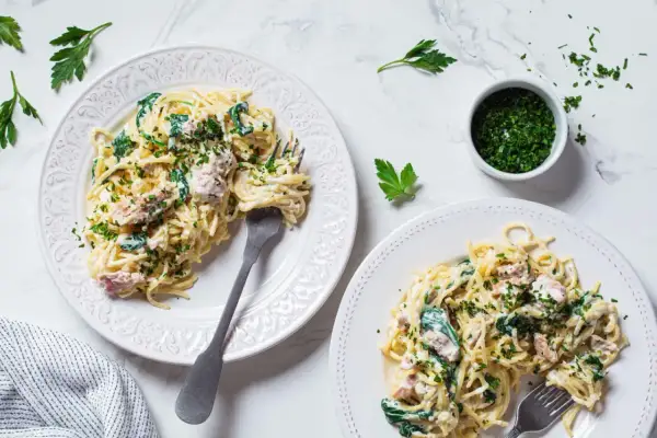 Dwa talerze spaghetti z kremowym sosem, szpinakiem i kawałkami tuńczyka z puszki, posypane natką pietruszki.