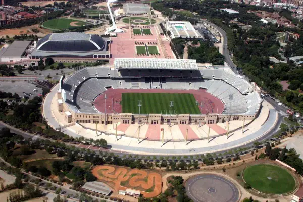 Widok z lotu ptaka na stadion olimpijski Montjuïc z zieloną murawą i trybunami. W pobliżu inne obiekty sportowe i tereny zielone.