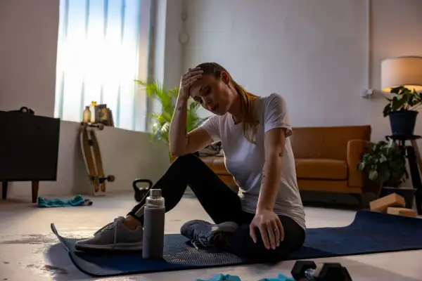 Erschöpfte Frau sitzt auf einer Matte, Hand an der Stirn. Vielleicht ein Fehler beim Muskelaufbau? Trainingsgeräte liegen herum.