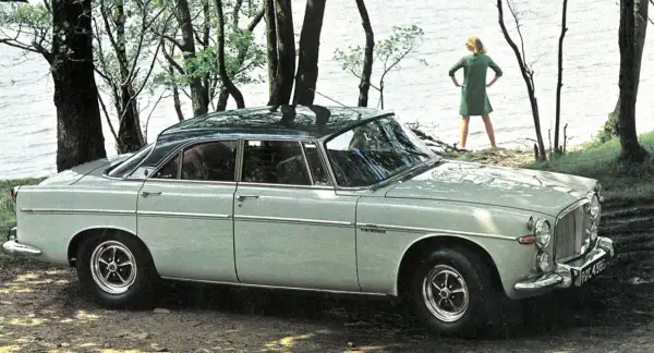 En ljusgrå Rover P5 står parkerad vid en sjö. En kvinna i grön klänning står vid vattenbrynet.