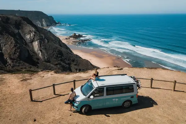 Ein Campervan steht auf einer Klippe mit Blick auf die atemberaubende portugiesische Natur und den Ozean. Zwei Personen genießen die Aussicht.