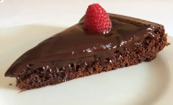Una fetta di gateau al cioccolato, lucida e golosa, con una fragola fresca in cima, su un piatto bianco.