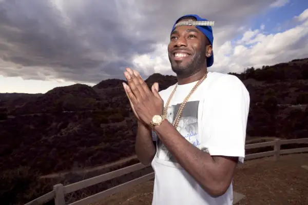 Dougie F. smiles and claps his hands, wearing a blue cap and gold chains, with a dramatic cloudy sky and hills behind him.