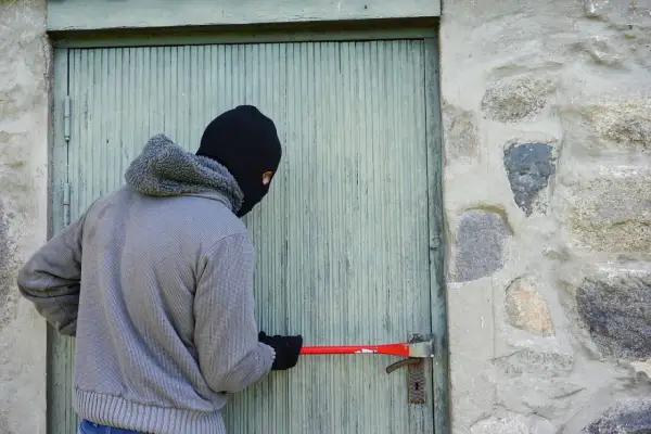 Un individu masqué tente de forcer une porte avec un pied-de-biche, acte relevant du cambriolage et du code pénal.