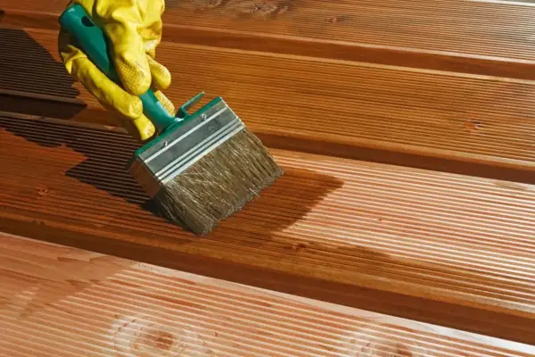 Ręka w żółtej rękawicy nakłada olej na drewniany taras. Zastanawiasz się, ile schnie olej do drewna?