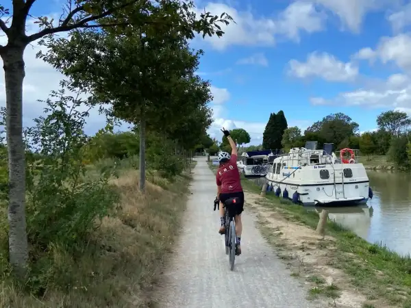 Ciclista pedaleando por el Canal du Midi, con barcos amarrados a un lado y árboles al otro.