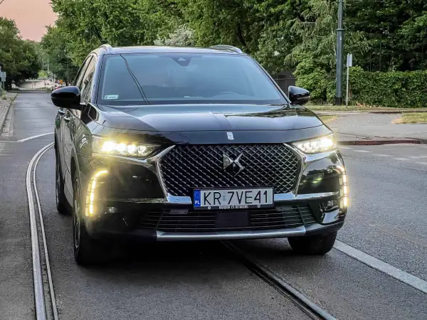 Czarny DS 7 z włączonymi światłami dziennymi stoi na torach tramwajowych.