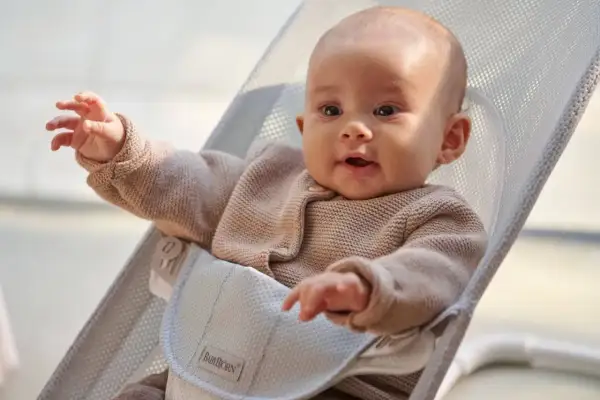 Uśmiechnięte niemowlę w beżowym sweterku siedzi w leżaczku BabyBjörn. Ten leżaczek jest idealny od pierwszych miesięcy życia dziecka.
