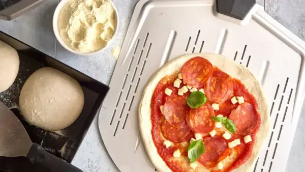 Pizzadeg recept: färdig pizzadeg, tomatsås, salami och ost på en pizzaspade. Bredvid en skål med mjöl och en form med pizzadeg.