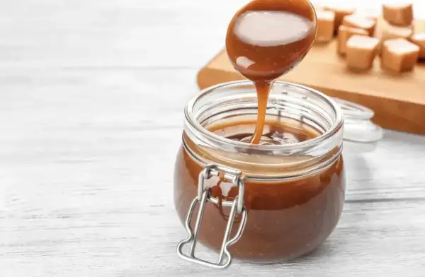 Une cuillère verse un coulis de caramel onctueux dans un bocal en verre. Des bonbons au caramel sont disposés sur une planche en bois en arrière-plan.