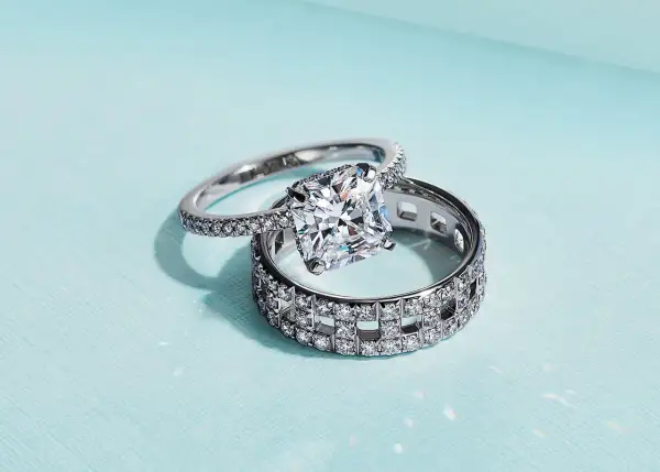Un anillo de compromiso con un gran diamante cuadrado reposa sobre un anillo de matrimonio con incrustaciones de diamantes, ambos de plata.