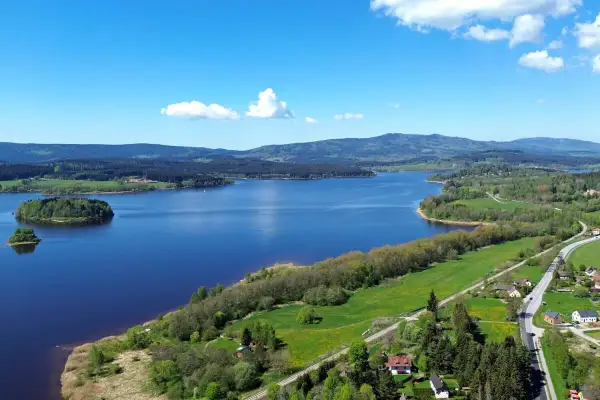 Weite Sicht auf den Lipno Stausee mit bewaldeten Hügeln, grünen Wiesen und kleinen Häusern am Ufer.