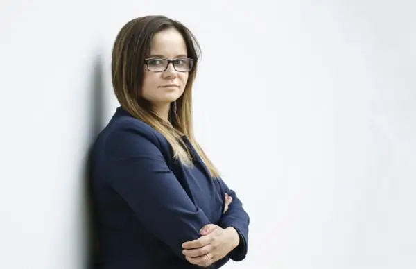 Kobieta w okularach i granatowej marynarce stoi oparta o ścianę, ze skrzyżowanymi rękami.
