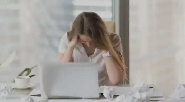 En kvinna med utmattningssyndrom känner yrsel och håller huvudet i händerna vid ett skrivbord med en laptop och papper.