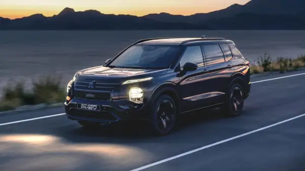 Ciemny Mitsubishi Outlander PHEV jedzie drogą o zachodzie słońca, z górami i jeziorem w tle.