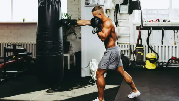 A man boxing with a heavy bag in a gym. He's wearing boxing gloves and shorts, focused on his training.